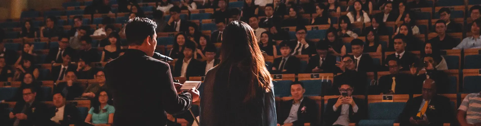 A man and woman's back faces the camera as they speak to an audience